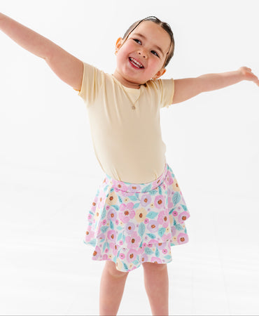 Child wearing a yellow shirt and floral skort with arms outstretched on a white background