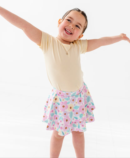 Child wearing a yellow shirt and floral skort with arms outstretched on a white background
