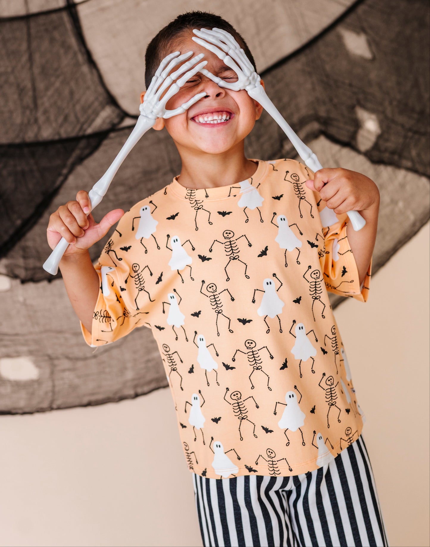 Child wearing a Halloween-themed shirt with skeleton and ghost designs, holding white skeleton hands over their eyes.