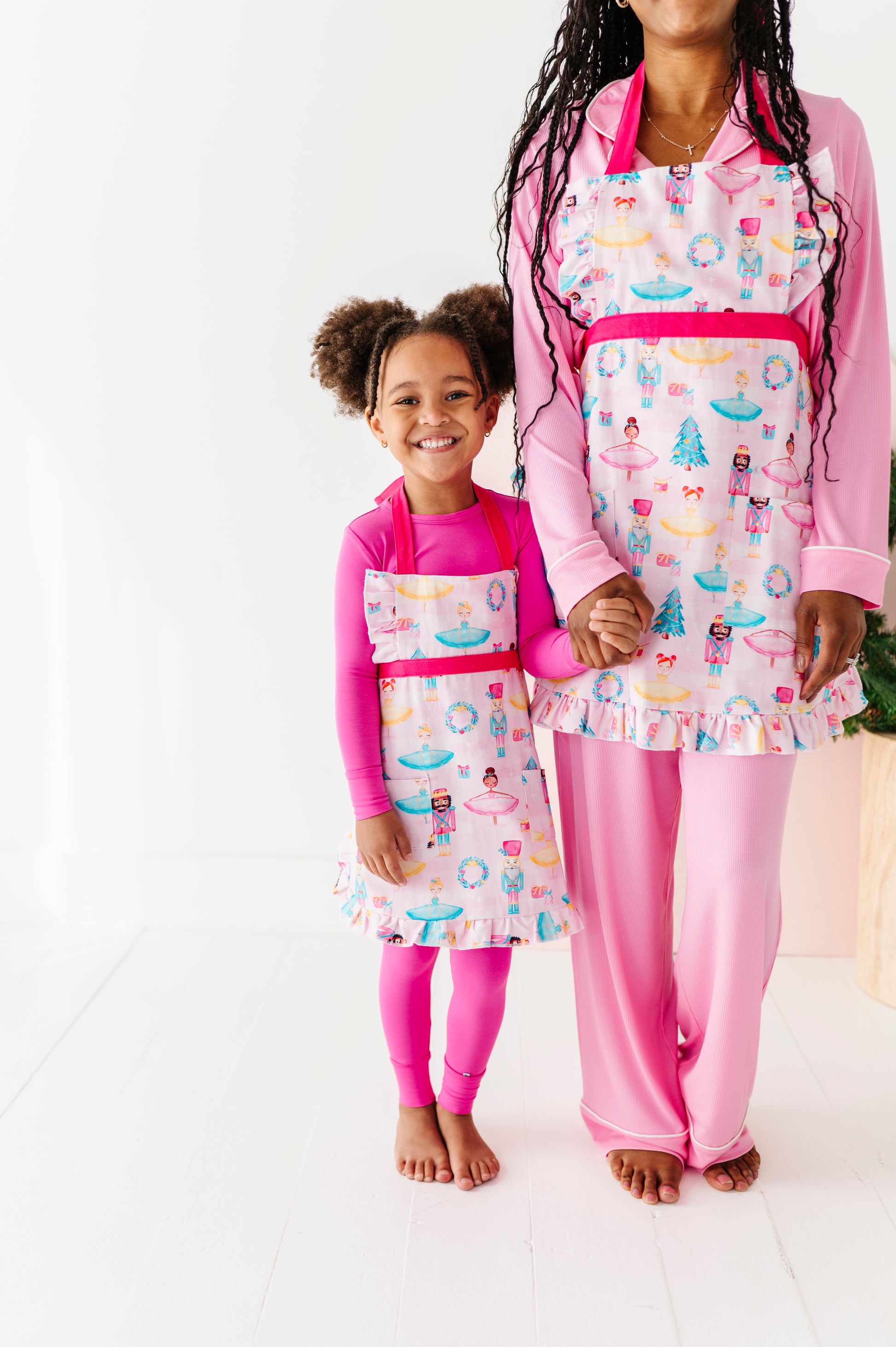A little girl and her mom wearing matching Christmas aprons featuring ballerinas, nutcrackers, trees, presents, and wreaths 