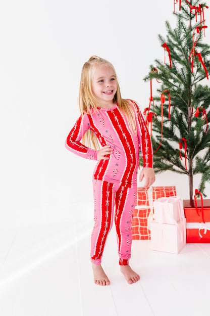 A young girl stands barefoot in a bright white room wearing a pink and red holiday-themed bamboo long-sleeve pajama set with candy cane and snowflake patterns. A decorated Christmas tree and wrapped gifts are visible in the background.