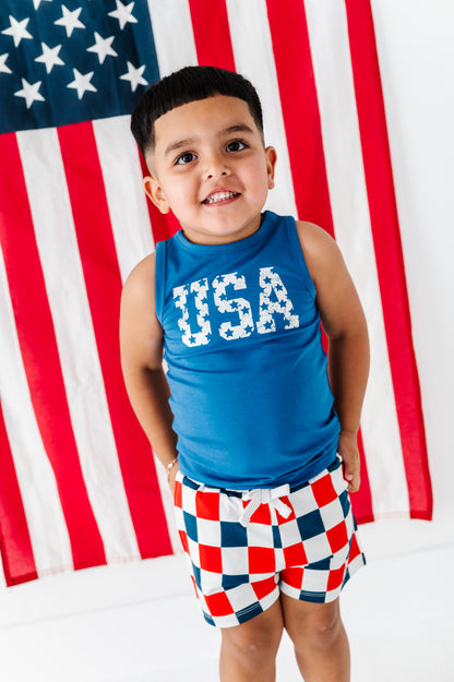 Child wearing a blue 'USA' bamboo tank and checkered shorts in front of an American flag