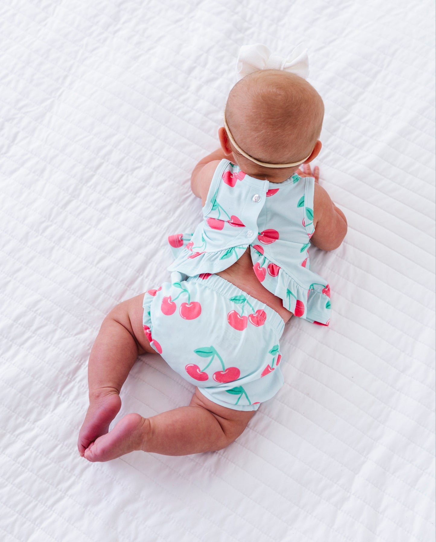 A baby lying on a white textured blanket, wearing a light blue cherry-print romper and matching headband with a white bow.