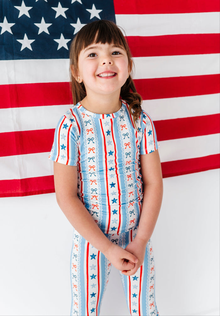 Child wearing a patriotic bamboo two-piece pajama set with red, white, and blue bows in front of an American flag