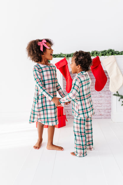 Two siblings, a girl wearing a bamboo nightgown in a classic Christmas plaid print and a boy wearing a matching bamboo lounge set, dancing in front of a fireplace 