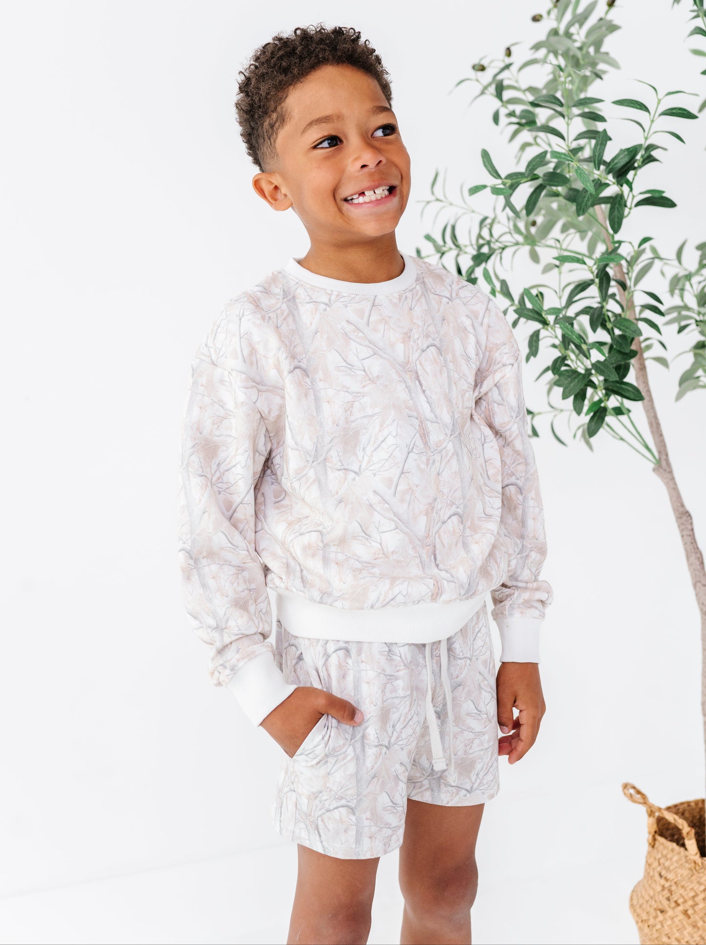 Child wearing a white patterned outfit standing next to a plant on a white background