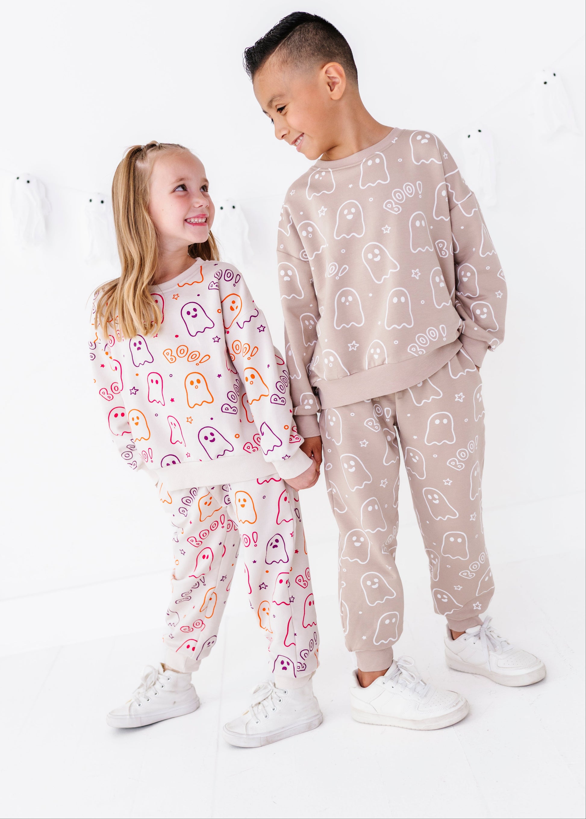 Two children modeling matching ghost-print sweatshirt and jogger sets in a bright white studio. The girl wears a white set with pink, orange, and purple ghosts, and the boy wears a beige set with white ghosts.