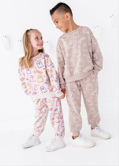 Two children modeling matching ghost-print sweatshirt and jogger sets in a bright white studio. The girl wears a white set with pink, orange, and purple ghosts, and the boy wears a beige set with white ghosts.