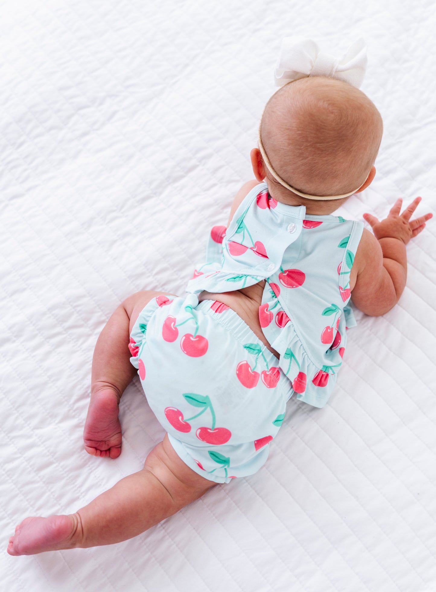 A baby lying on a white quilted blanket, viewed from behind, wearing a light blue sleeveless outfit with a cherry print and a matching bow headband.