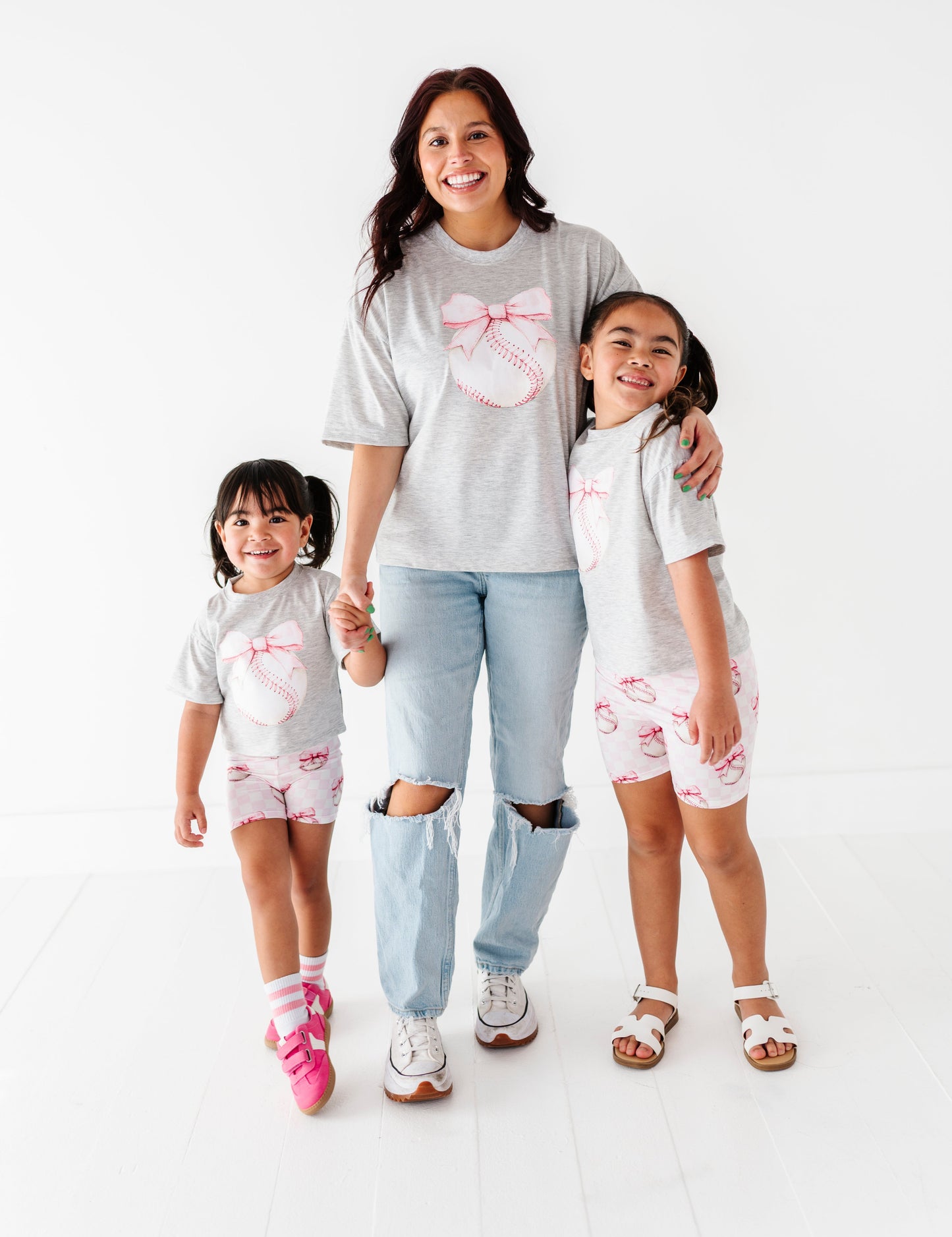 Woman and two children wearing matching outfits with a pink bow baseball design on a white background