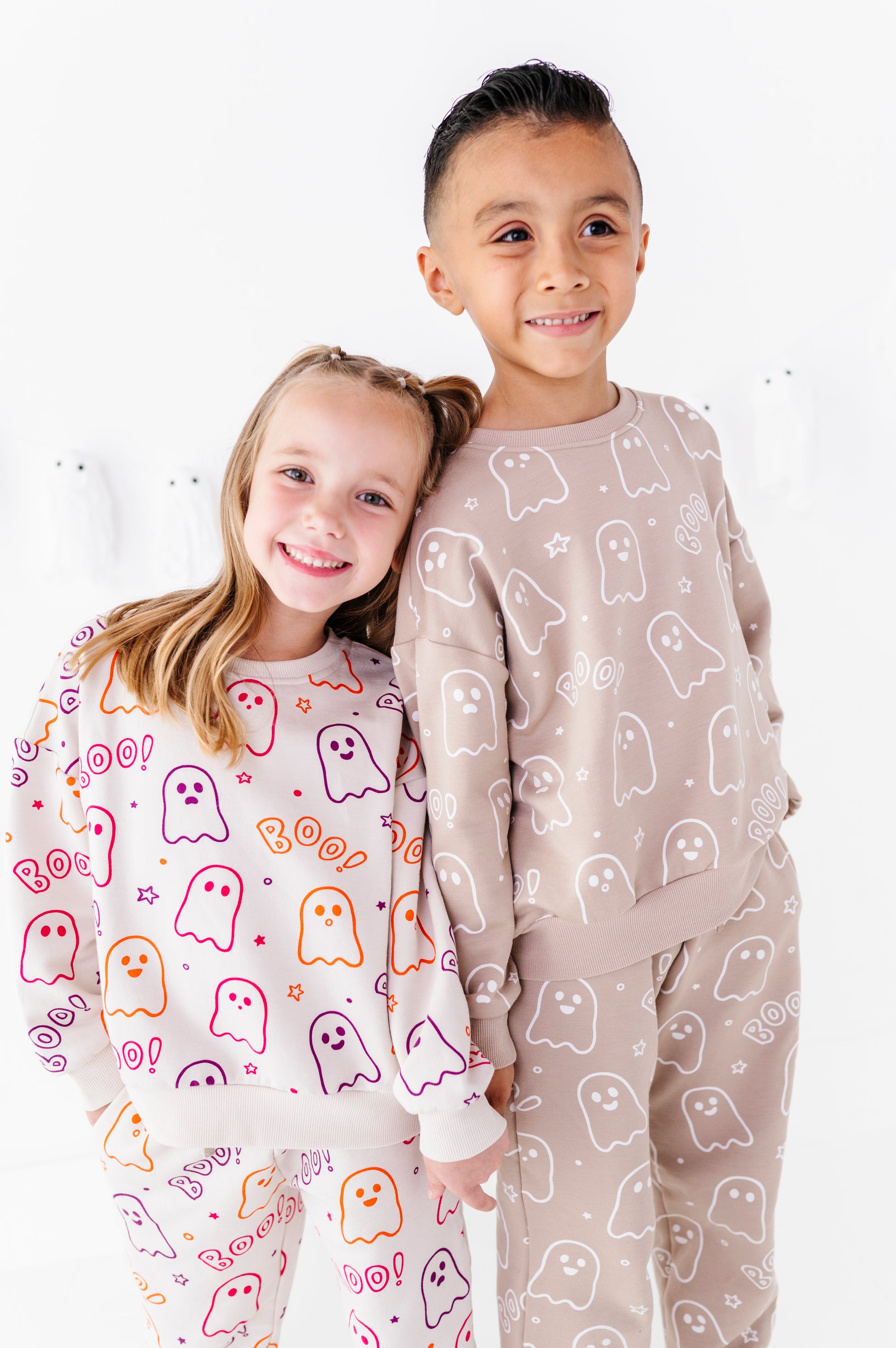 Two children wearing matching ghost-print jogger sets, standing against a clean white background. The girl wears a white set with colorful orange, pink, and purple ghosts, and the boy wears a light beige set with white ghosts.