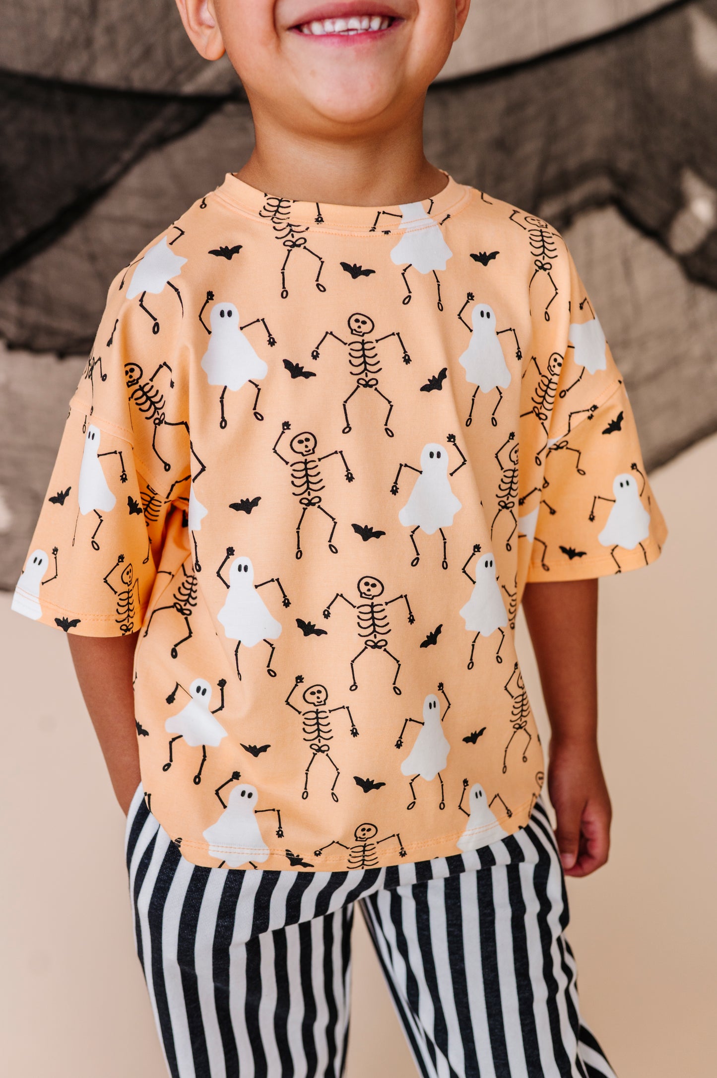 Child wearing a Halloween-themed shirt with ghosts and skeletons on an orange background.