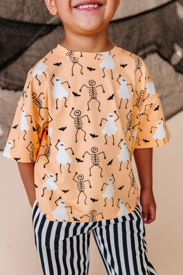 Child wearing a Halloween-themed shirt with ghosts and skeletons on an orange background.