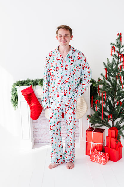 A man wearing a light blue Christmas bamboo pajama set with a Santa Claus print stands barefoot in a brightly lit room decorated for Christmas, with a fireplace, red stocking, and decorated tree nearby.