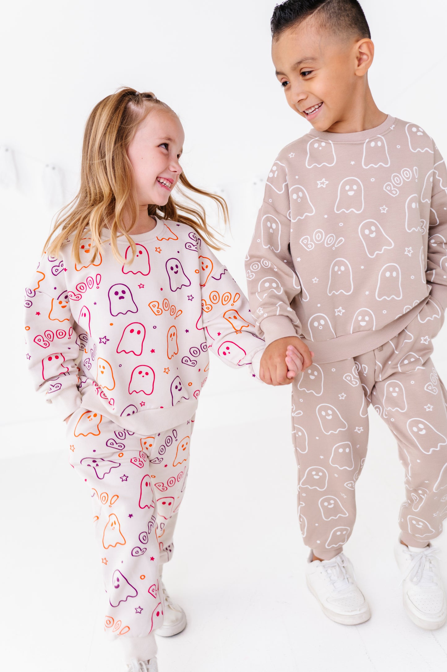 Two children wearing matching ghost-print jogger sets, standing and holding hands in a bright white room. The girl wears a white set with colorful orange, pink, and purple ghost outlines, and the boy wears a beige set with white ghost outlines.