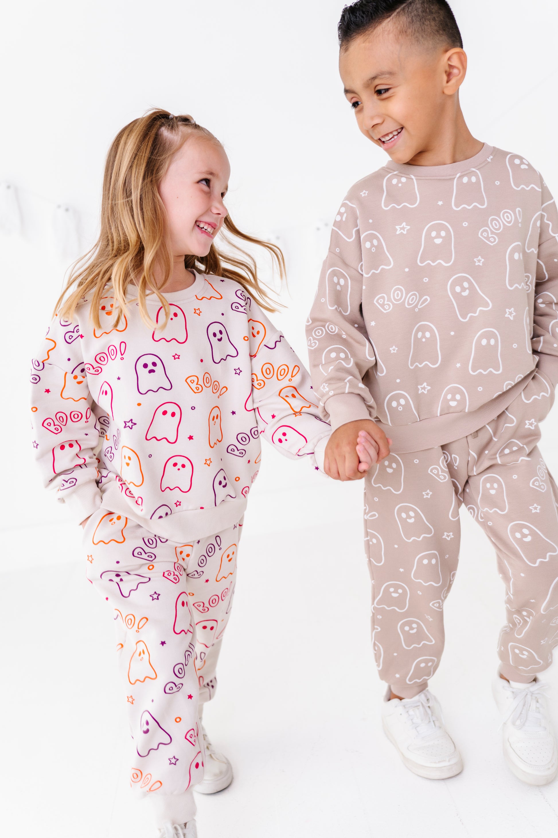Two children wearing matching ghost-print jogger sets, standing and holding hands in a bright white room. The girl wears a white set with colorful orange, pink, and purple ghost outlines, and the boy wears a beige set with white ghost outlines.