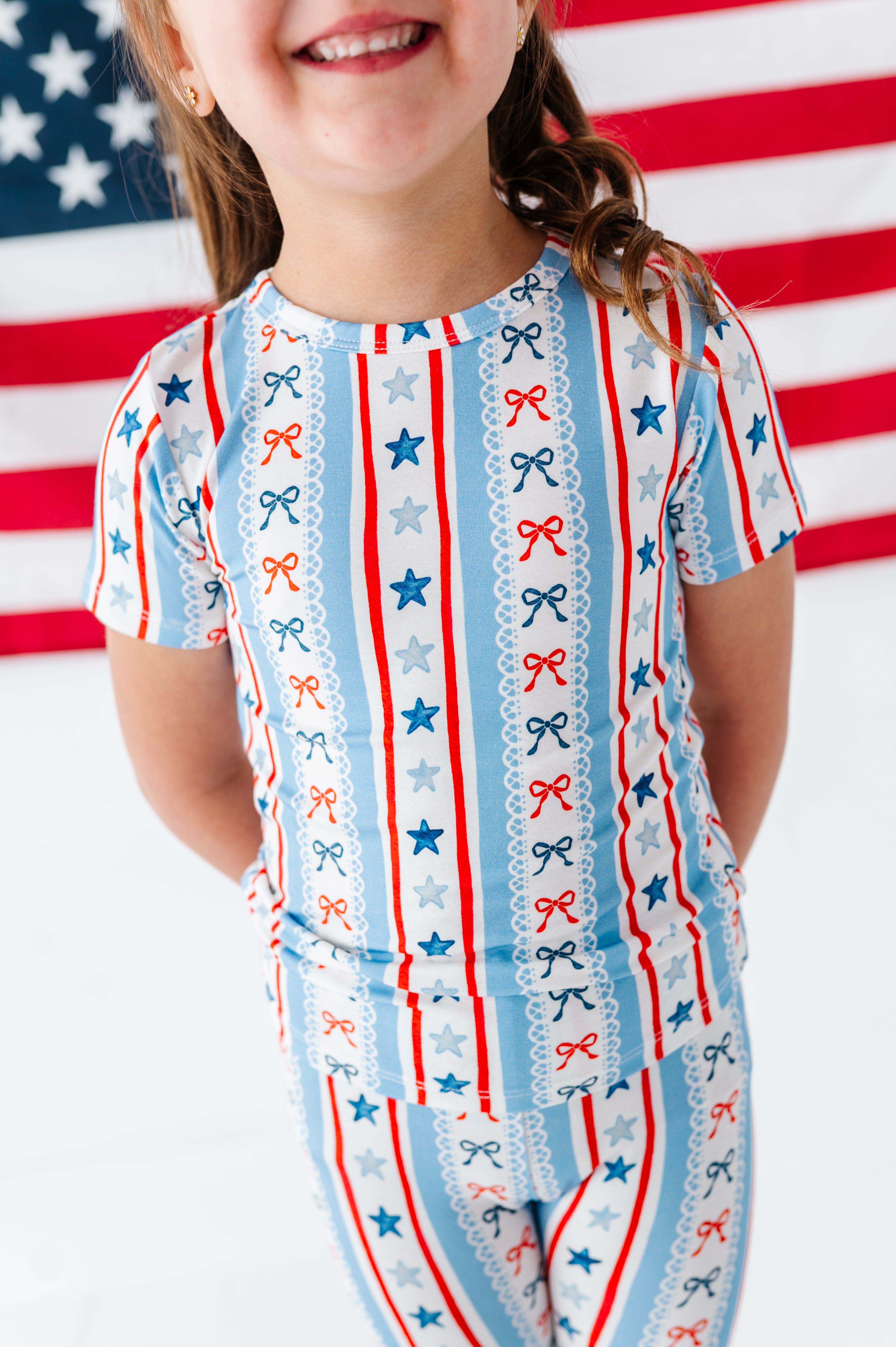 Little girl wearing a patriotic-themed bamboo two-piece set pajama outfit with stars and stripes in front of an American flag.