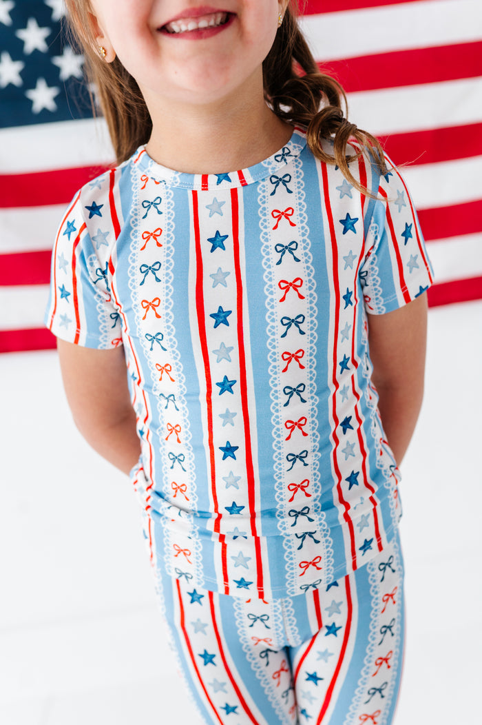 Little girl wearing a patriotic-themed bamboo two-piece set pajama outfit with stars and stripes in front of an American flag.