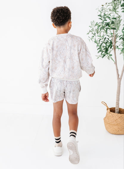 Child wearing a camo outfit standing in a minimalistic room with a plant.
