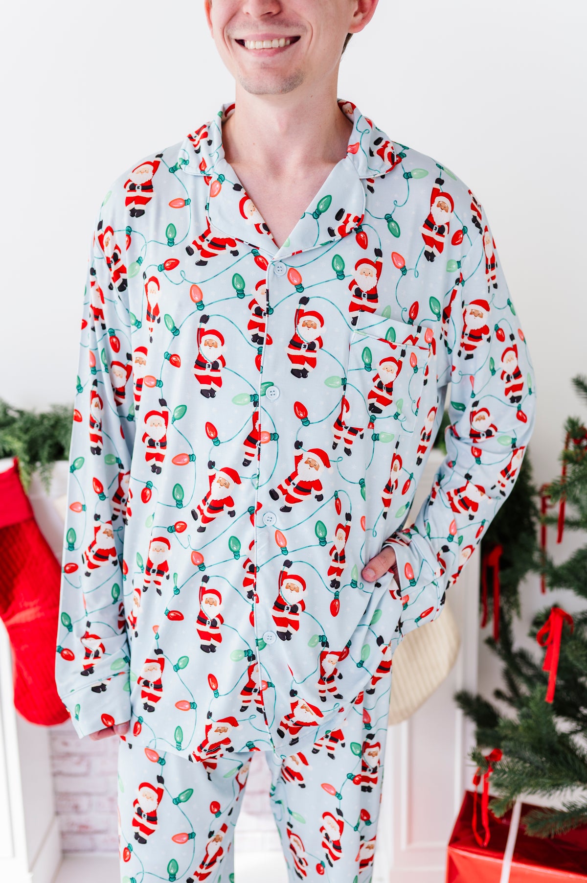 A man wearing a light blue long-sleeve bamboo pajama set with a festive Christmas print of Santa Claus, stockings, and a Christmas tree, standing in a holiday-themed room.