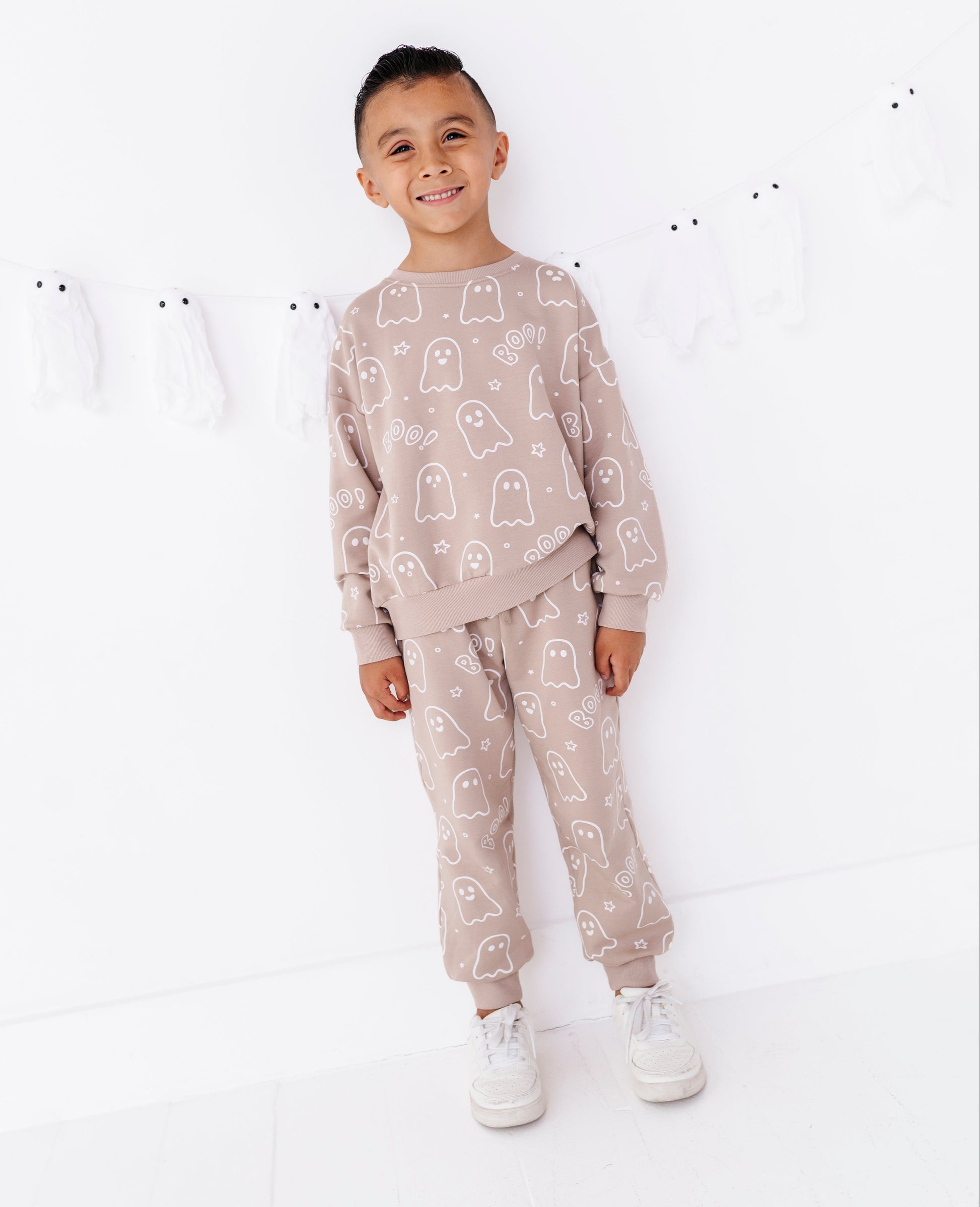 A young boy stands smiling in a matching beige pajama set with a white ghost pattern, featuring a long-sleeve top and jogger pants, paired with white sneakers against a white background.