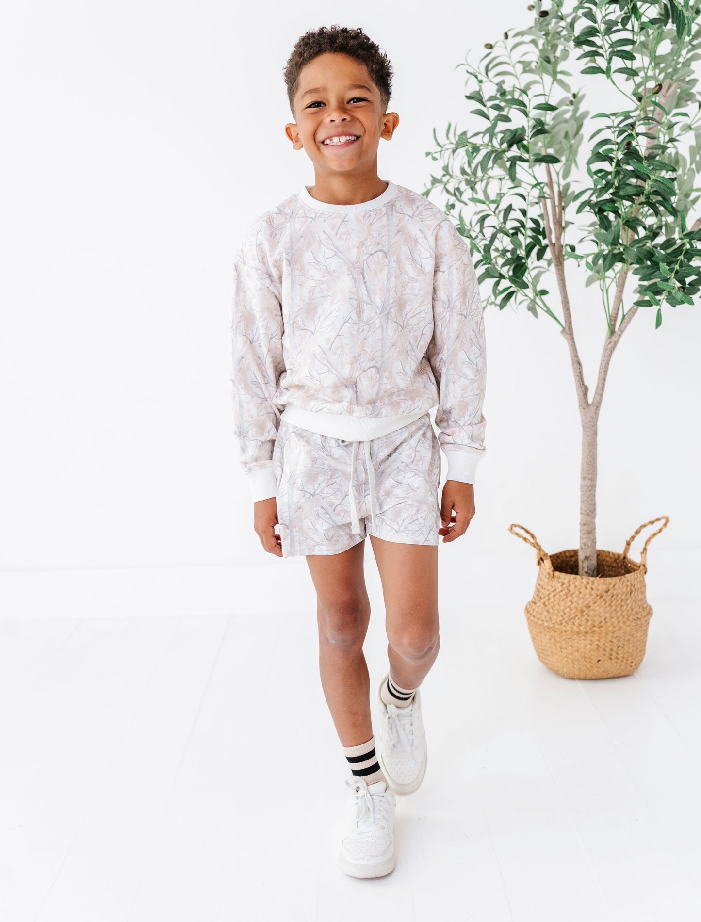 Child wearing a light-colored camo pattern outfit standing next to a potted plant on a white background