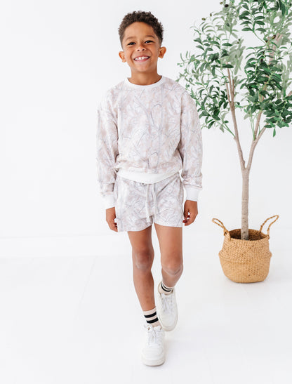 Child wearing a light-colored camo pattern outfit standing next to a potted plant on a white background