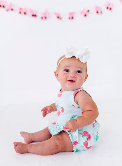 A baby girl sitting on a white studio floor wearing a light blue cherry print romper and a white bow headband.