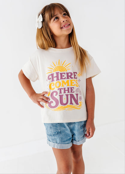 Young girl wearing a  t-shirt with 'Here Comes the Sun' text and sun graphic on a white background