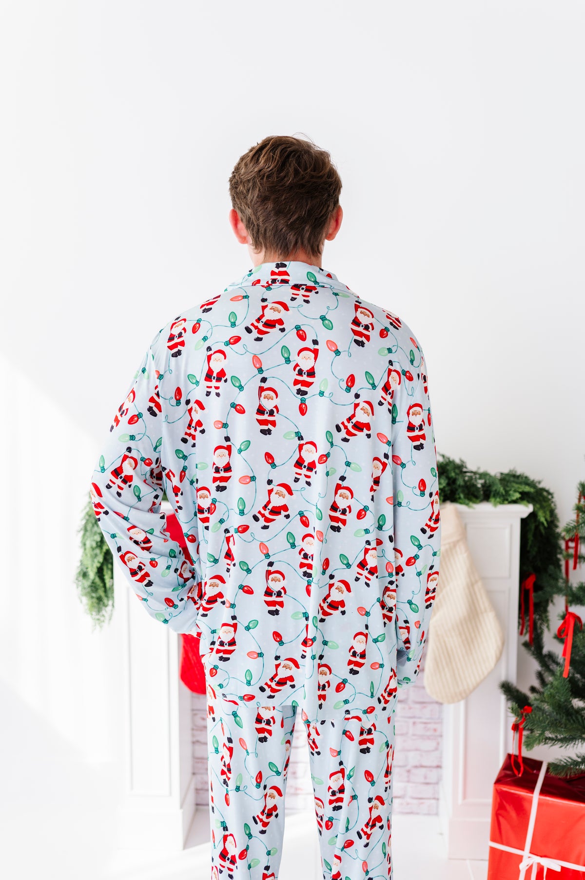 A man viewed from behind wearing a light blue Christmas-themed bamboo pajama set with a repeating Santa Claus pattern, standing in a brightly lit room with holiday decorations.