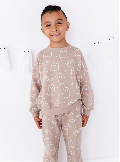 A young boy wearing a matching beige and white ghost-patterned sweatshirt and sweatpants set, standing against a white wall with white ghost decorations.