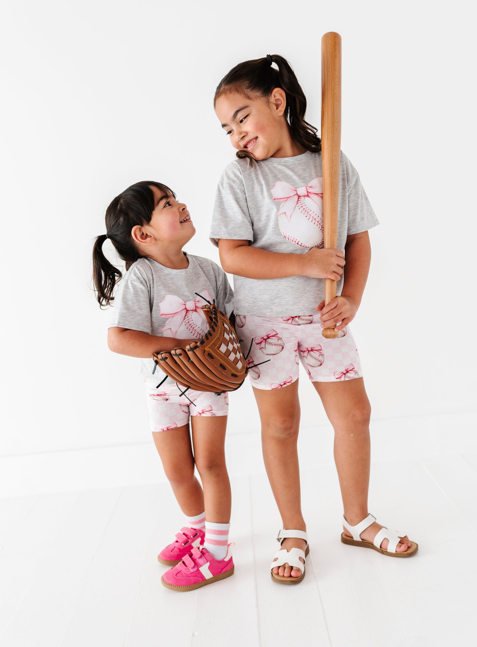 Two young girls in matching baseball outfits with a bat and glove on a white background