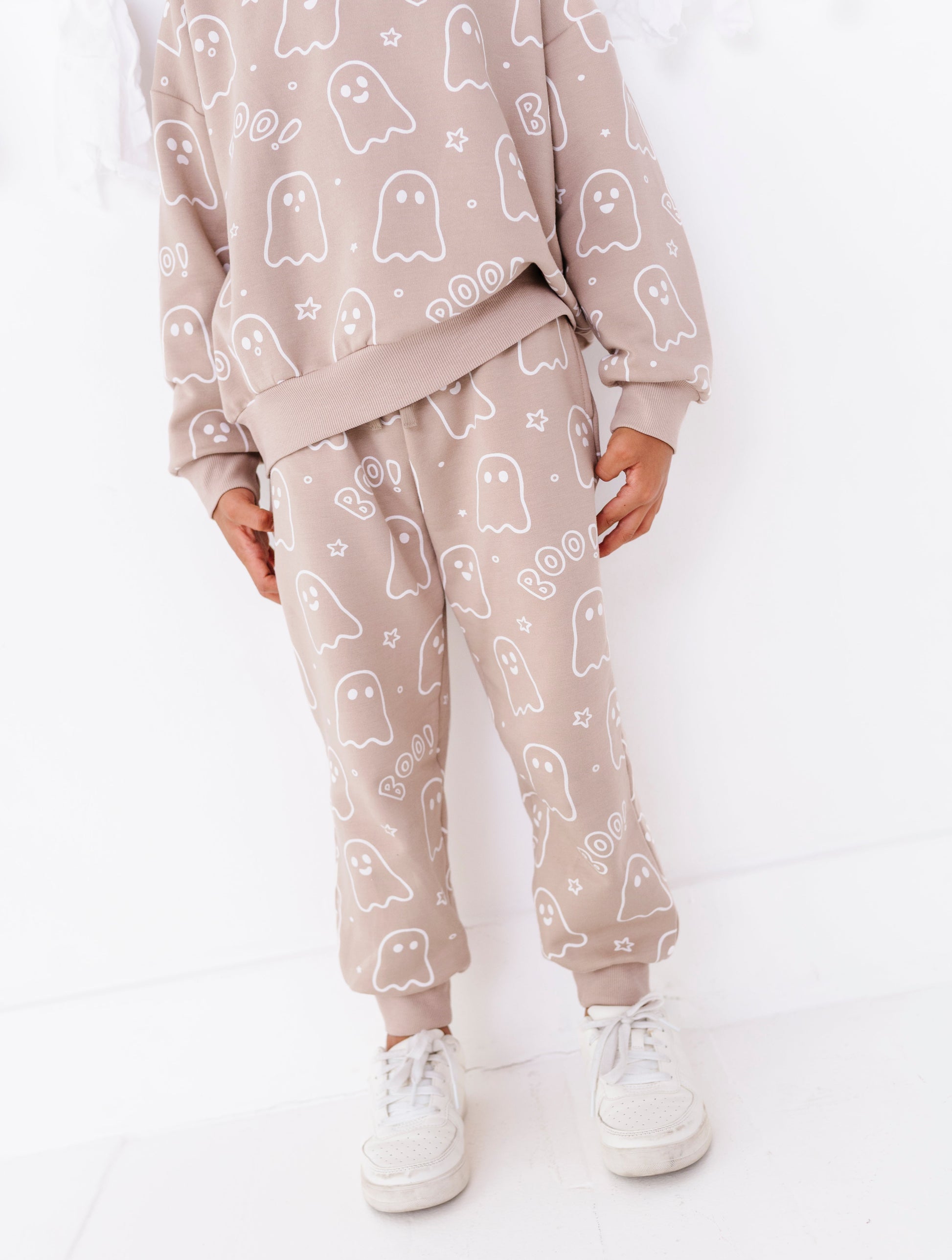 A child wearing a matching beige pajama set with a white ghost pattern, featuring a sweatshirt and jogger pants, paired with white sneakers.