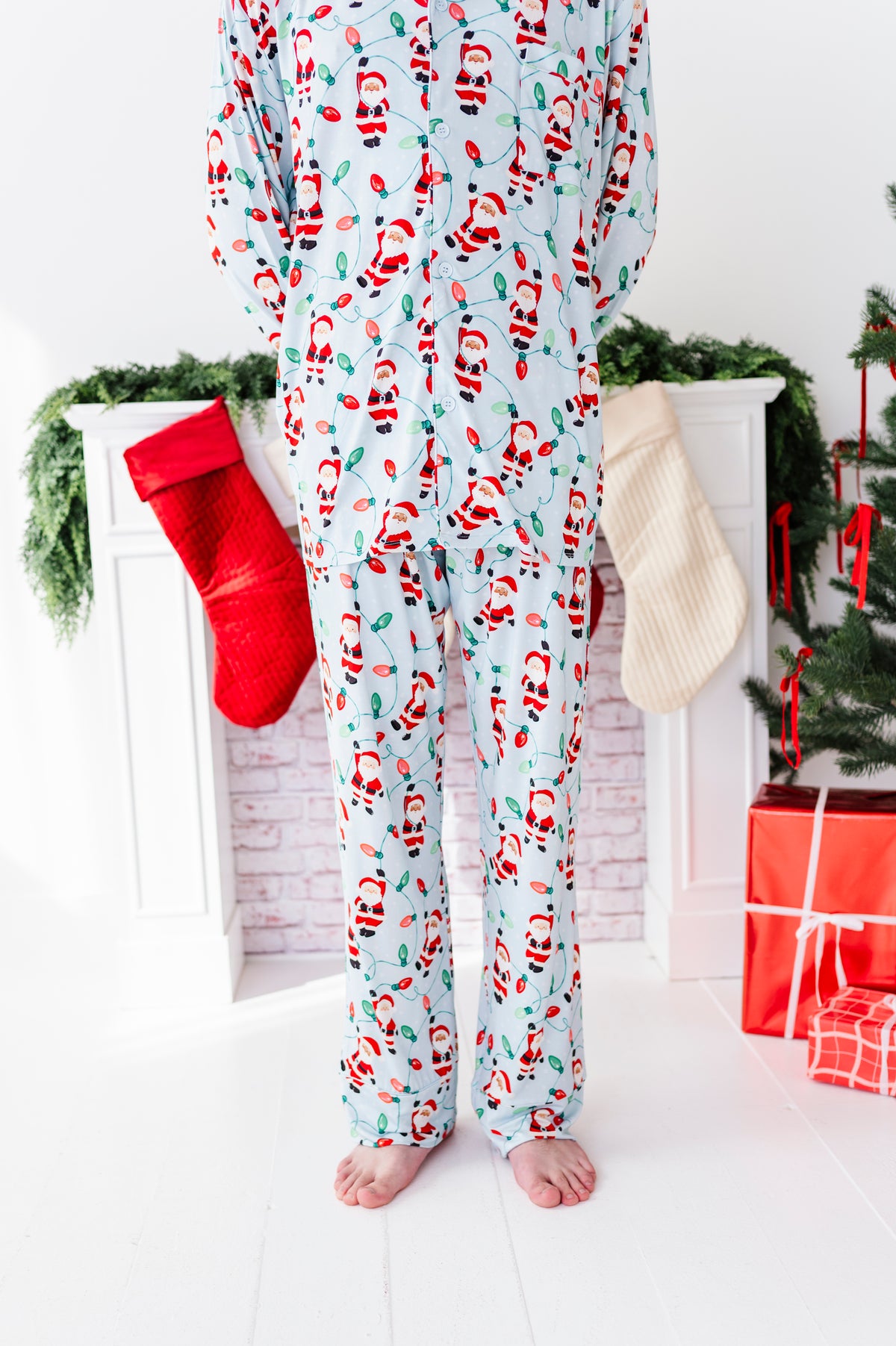 A person wearing light blue Christmas pajama bamboo set with a Santa Claus print, standing barefoot in front of a white fireplace decorated with stockings, greenery, and wrapped gifts.