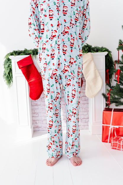 A person wearing light blue Christmas pajama bamboo set with a Santa Claus print, standing barefoot in front of a white fireplace decorated with stockings, greenery, and wrapped gifts.