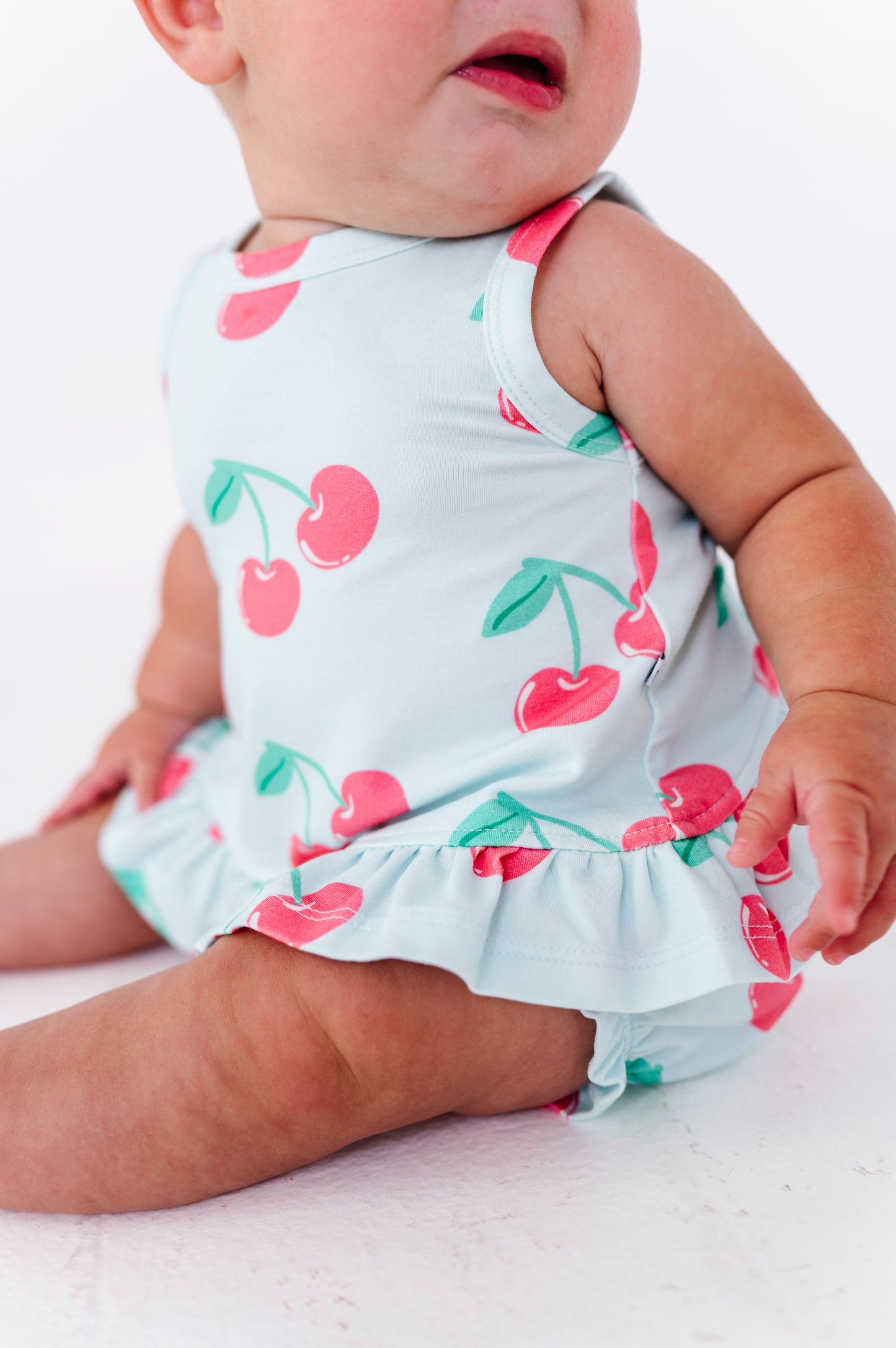 A close-up of a baby wearing a light blue sleeveless outfit with a pink and green cherry print, sitting against a white background.
