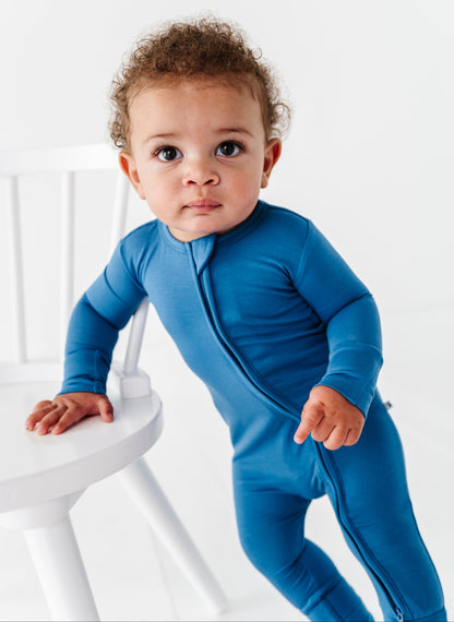 Baby in a blue bamboo zip romper standing on a white chair against a white background