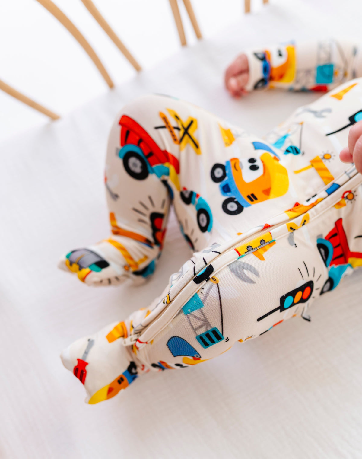 A baby wearing a cream-colored footed sleeper with a colorful construction vehicle print, lying on a white mattress in a wooden crib.