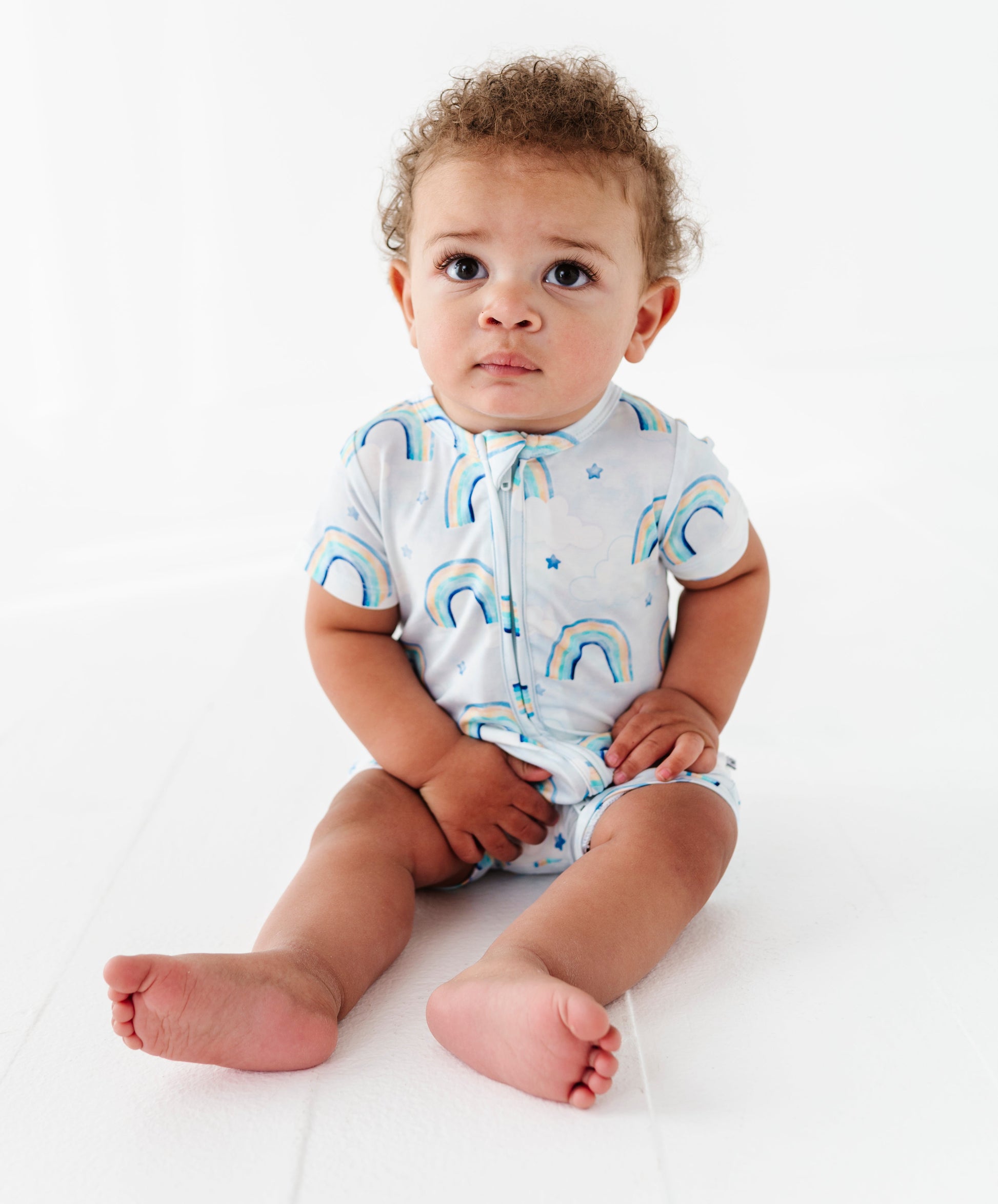Baby wearing a white bamboo shortie romper with blue watercolor rainbow patterns on a white background