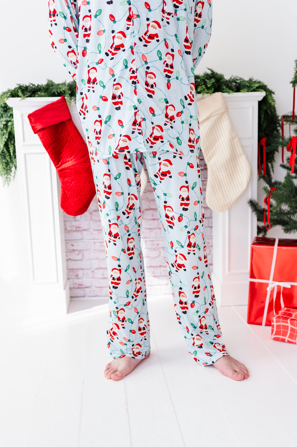 A person wearing light blue Christmas bamboo pajama pants with a Santa Claus print, standing barefoot in front of a white fireplace mantel decorated with a red stocking, green garland, and wrapped presents.