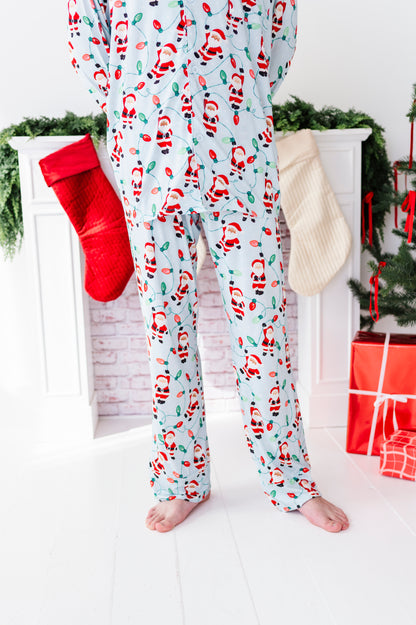 A person wearing light blue Christmas bamboo pajama pants with a Santa Claus print, standing barefoot in front of a white fireplace mantel decorated with a red stocking, green garland, and wrapped presents.