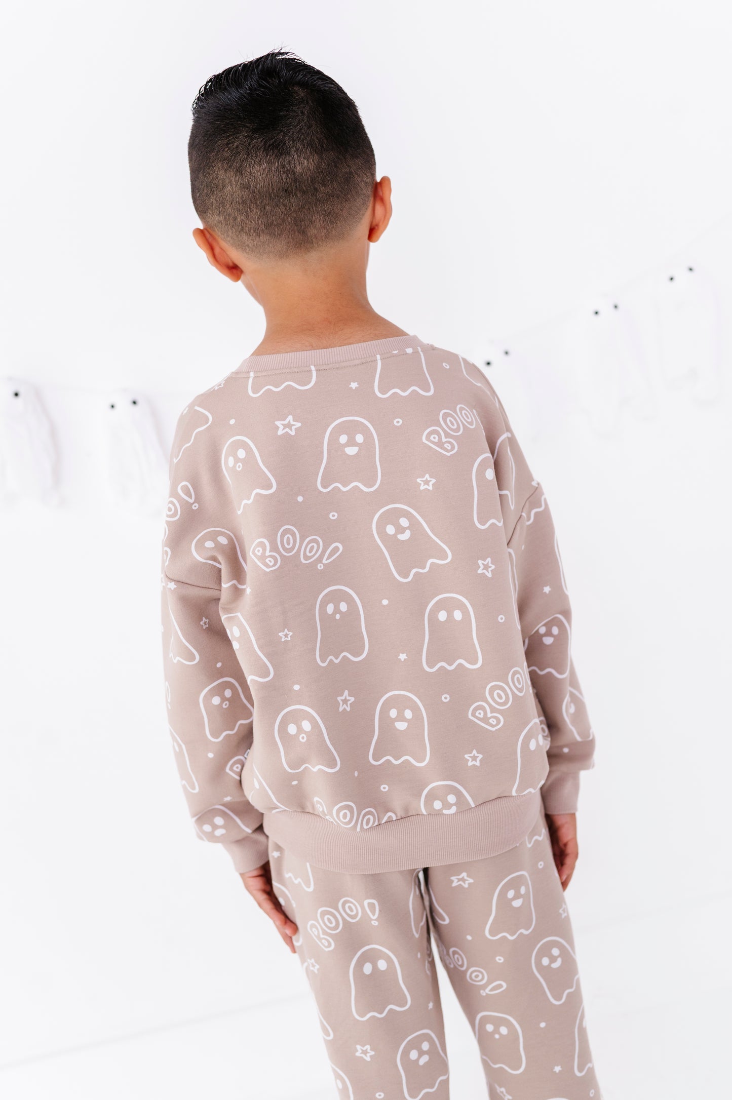A child viewed from behind wearing a matching light beige pajama set with a white ghost and 'Boo' pattern, set against a clean white background.