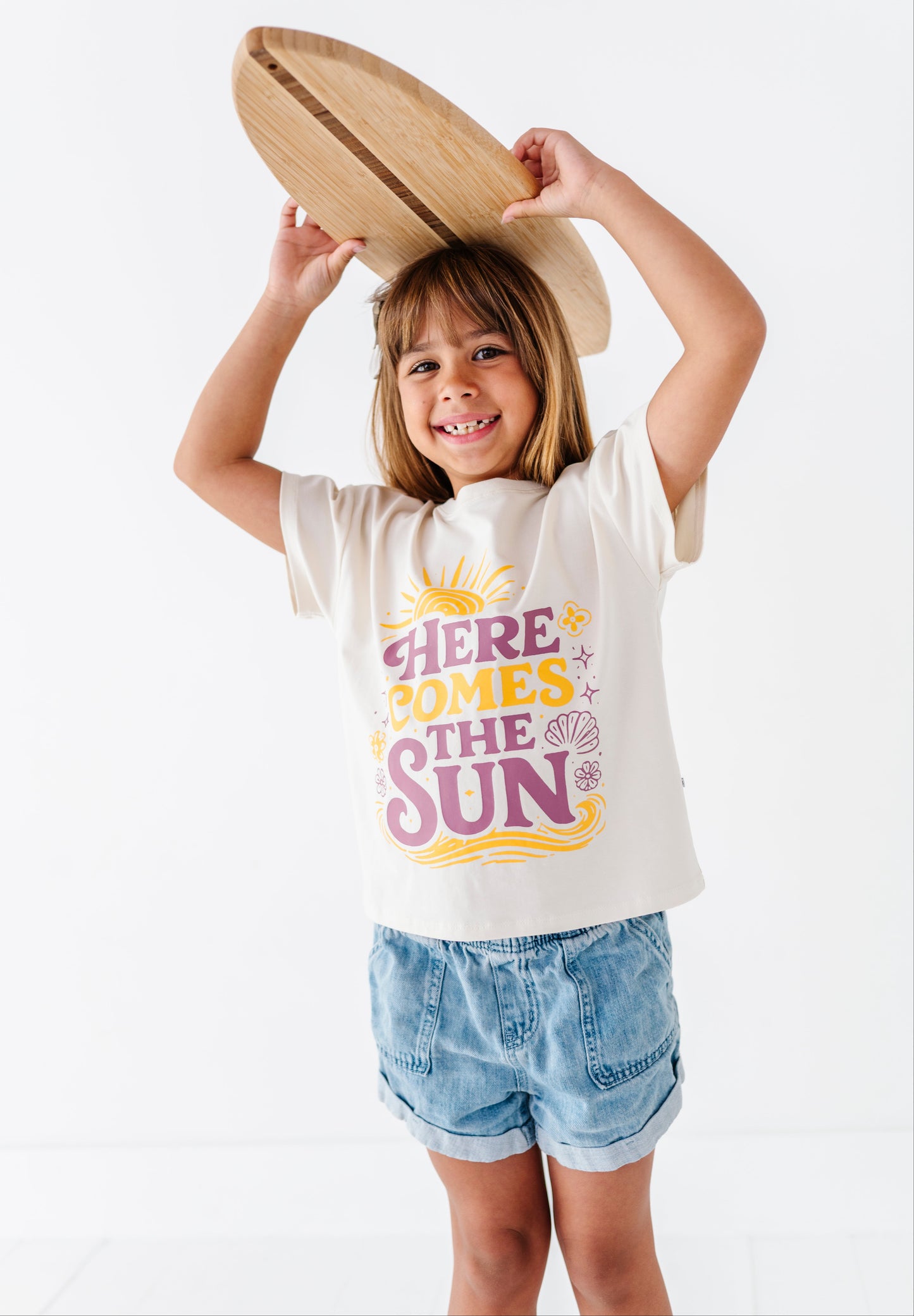 Child wearing a 'Here Comes the Sun' t-shirt holding a wooden surfboard on a white background