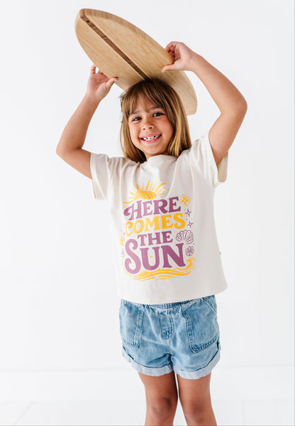 Child wearing a 'Here Comes the Sun' t-shirt holding a wooden surfboard on a white background