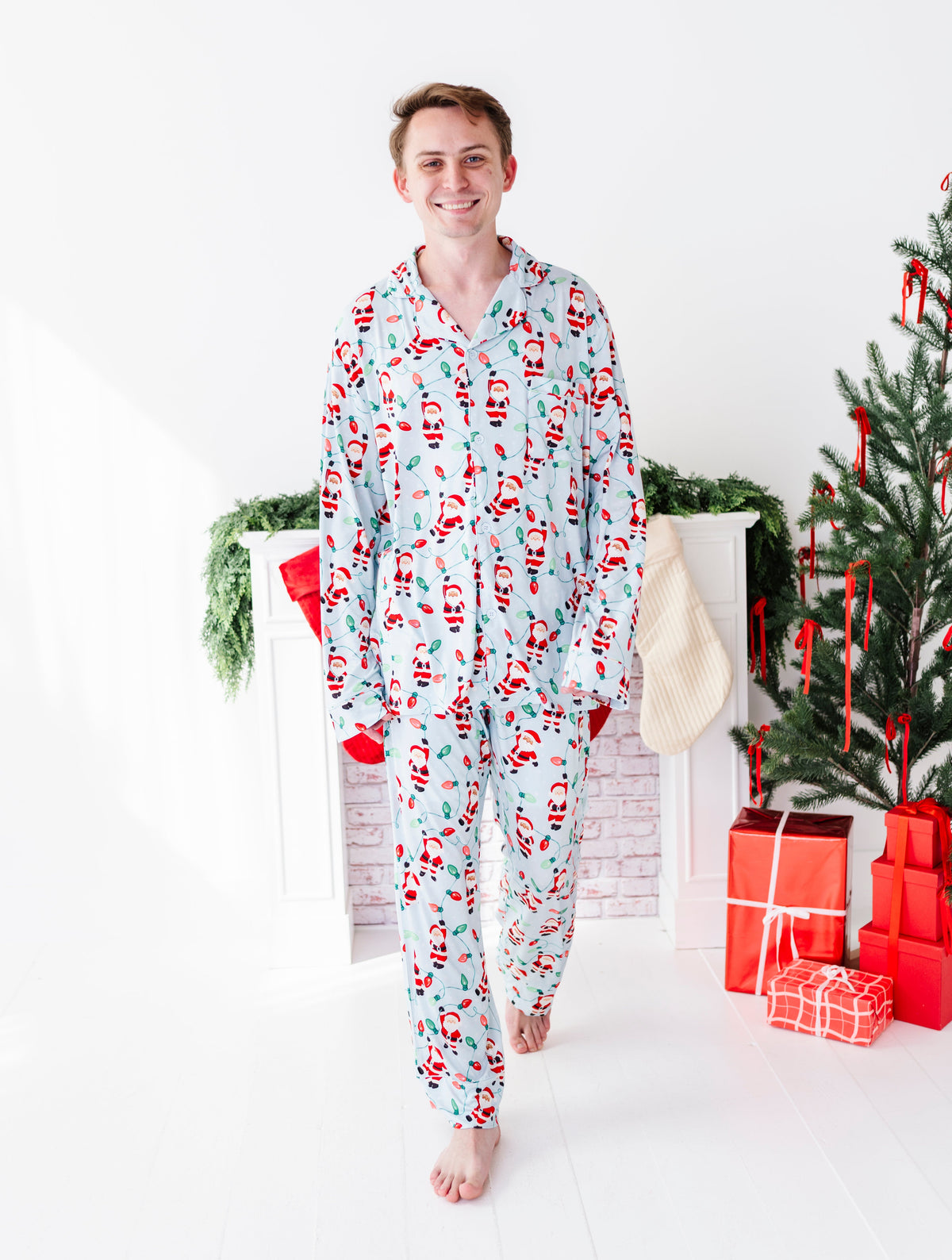 A man wearing a light blue Christmas-themed bamboo pajama set, standing in a brightly lit room decorated with a Christmas tree, stockings, and wrapped gifts.