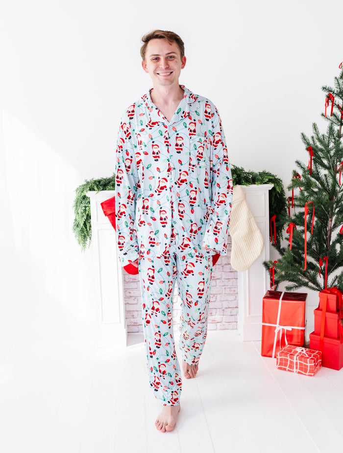 A man wearing a light blue Christmas-themed bamboo pajama set, standing in a brightly lit room decorated with a Christmas tree, stockings, and wrapped gifts.