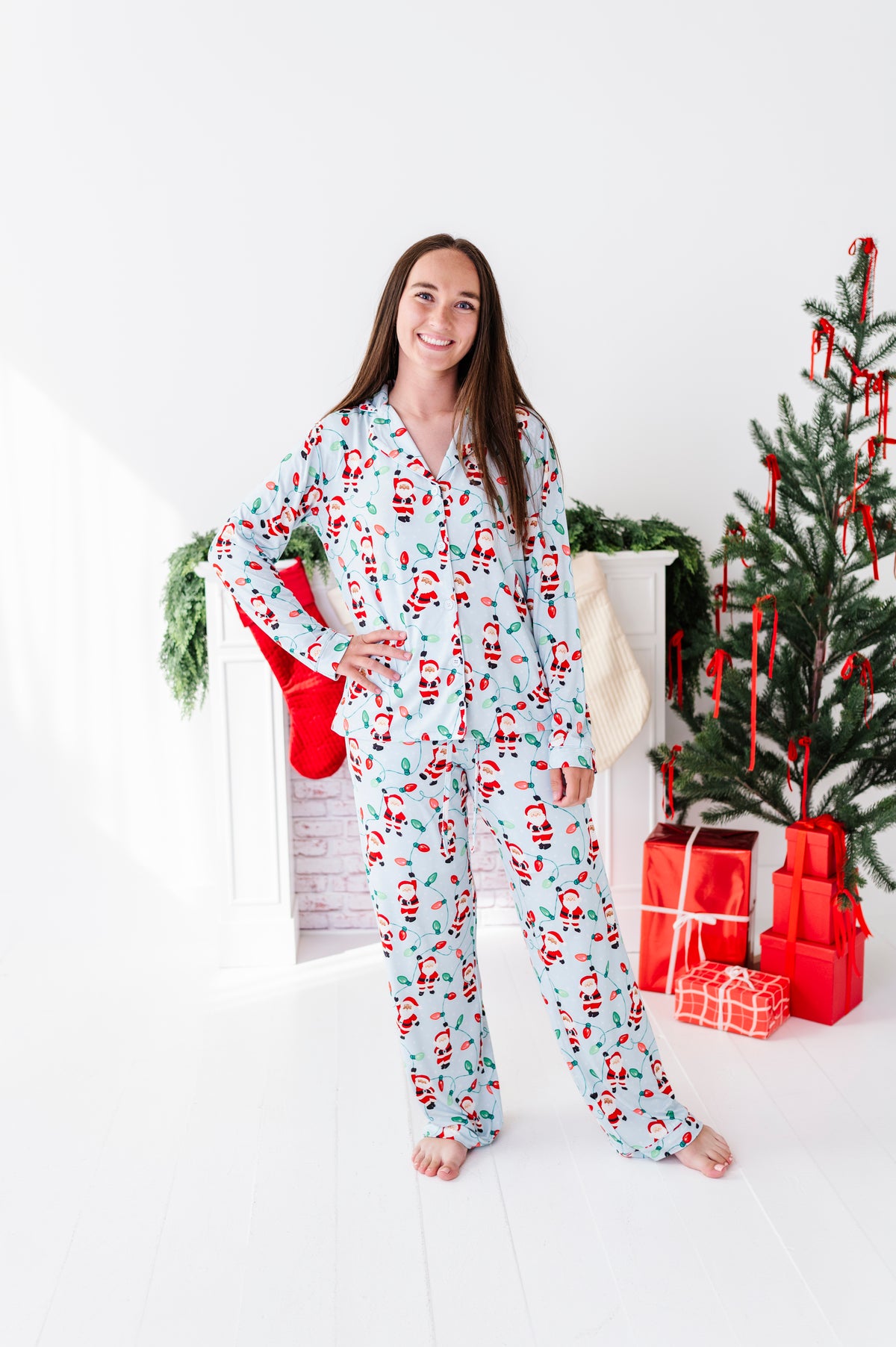 A woman wearing a light blue long-sleeve bamboo pajama set with a red and green Santa Claus and Christmas tree print stands barefoot in a bright white room decorated for Christmas, with a tree, stockings, and wrapped gifts in the background.