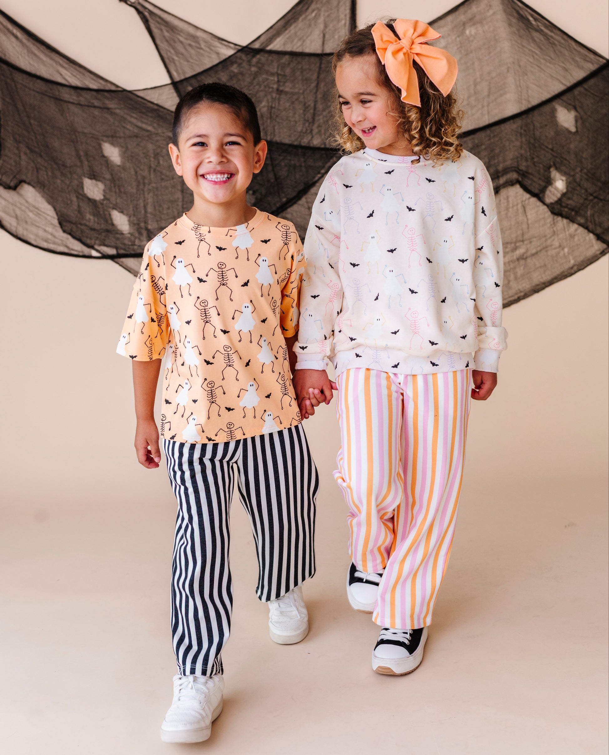 Two children holding hands in front of a decorative net, wearing halloween patterned outfits.
