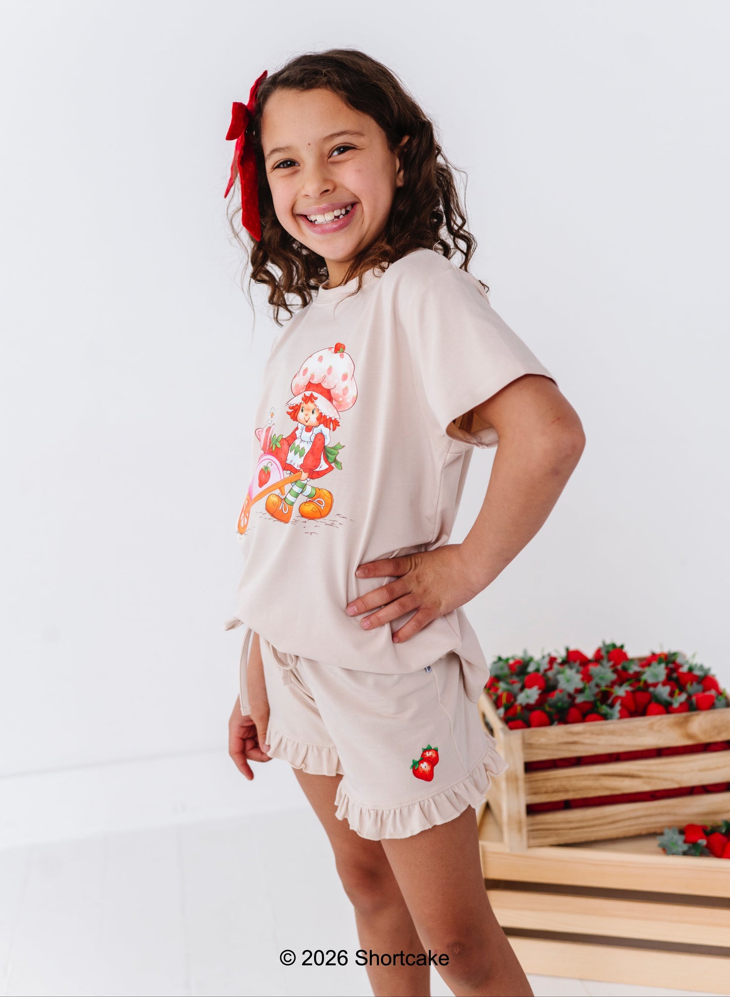 Young girl wearing a beige t-shirt with a Strawberry Shortcake™ design and matching, standing against a white background.