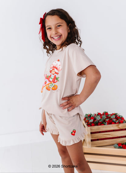 Young girl wearing a beige t-shirt with a Strawberry Shortcake™ design and matching, standing against a white background.